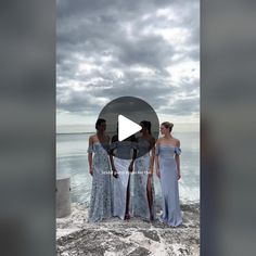 three women in dresses standing on rocks near the ocean with cloudy sky behind them and an overcast sky