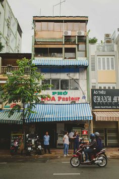 two people on a motorcycle in front of a building with blue and white awnings