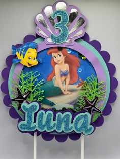 the little mermaid cake topper is decorated with blue and purple icing, under an ocean scene