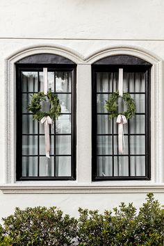 two wreaths on the windows of a white building with trees in front of them