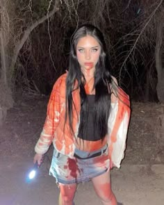 a woman with long black hair and red makeup is walking through the woods at night