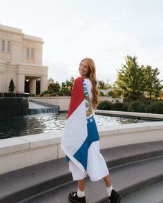 a woman standing on the steps in front of a fountain wearing a flag shirt and white skirt