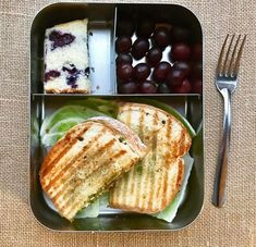 a lunch box with grapes, cake and sandwich on it next to a knife and fork