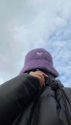 a woman wearing a purple hat and black jacket looking up at the sky with clouds in the background