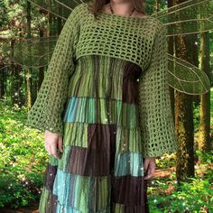 a woman standing in the woods wearing a green and brown crochet dress with long sleeves