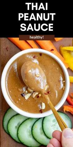 a hand holding a spoon over a bowl of peanut sauce with cucumbers and carrots in the background