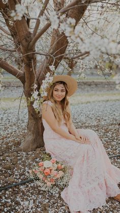 a woman wearing a hat sitting under a tree with flowers on the ground in front of her