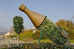 a sculpture of a large bottle with flowers on it's side in the middle of a flower bed