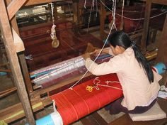 a woman sitting on the floor working on some kind of weaving machine with red yarn