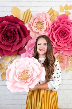 a woman holding large paper flowers in front of a wall with pink and red flowers