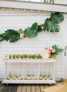 a white table topped with lots of bottles and glasses filled with drinks next to a green plant