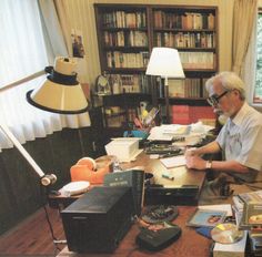 an older man sitting at a desk in front of a lamp and bookshelf