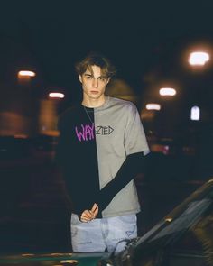 a young man standing in front of a car wearing a gray shirt and black sleeves