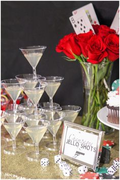 a table topped with lots of glasses filled with drinks and cupcakes next to a vase full of red roses