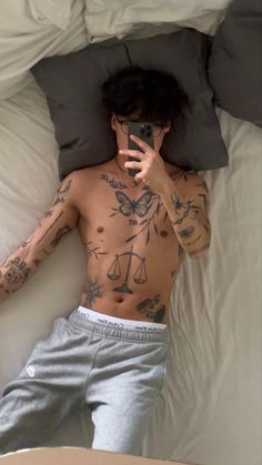 a man laying in bed with tattoos on his body and holding a cell phone up to his face