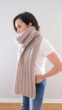 a woman wearing a knitted scarf and jeans stands in front of a white wall