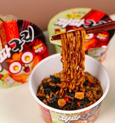 the noodles are being lifted by chopsticks from an ice cream cup with other snacks in the background