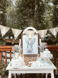 a white table topped with cakes and cupcakes next to a forest filled with trees