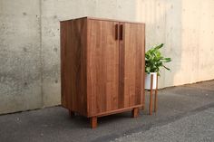 a wooden cabinet sitting next to a plant in a white vase on the side of a building