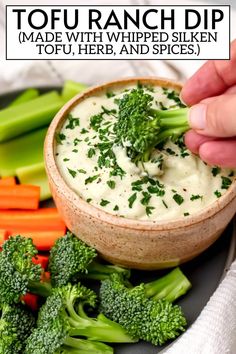 a person dipping dip into a bowl with broccoli, carrots and celery