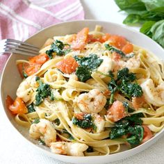 a white bowl filled with pasta and shrimp on top of a table next to basil leaves