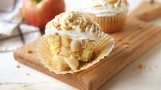 two cupcakes with white frosting and nuts on a cutting board next to an apple