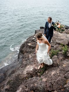 the bride and groom are walking along the rocks by the water