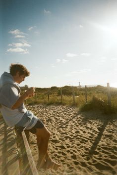 a man sitting on top of a wooden bench next to a sandy beach under a blue sky