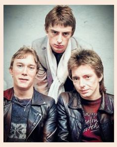 three young men are posing for a photo in front of a gray wall and one is wearing a black leather jacket