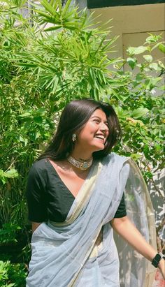 a woman in a sari smiles as she walks by some trees and bushes on a sunny day