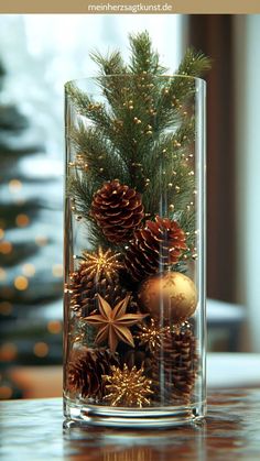 a glass vase filled with pine cones and other decorations on top of a table next to a christmas tree