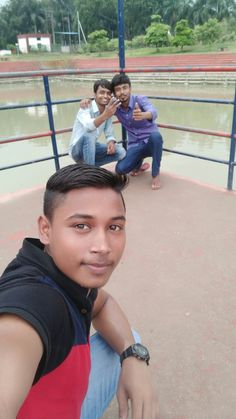 three men sitting on a bench in front of a pond and one man taking a selfie with his cell phone