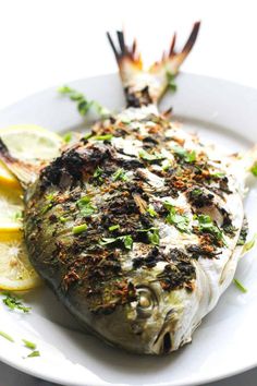 a white plate topped with a fish covered in herbs and lemon wedges next to a fork