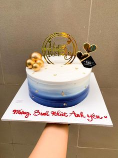 a white cake with blue and gold frosting sitting on top of a table next to a person's hand