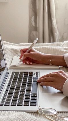 a woman writing on a notebook while using a laptop computer in her bed with eyeglasses