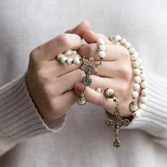 a woman holding a rosary and wearing a cross bracelet with white beads on her hands