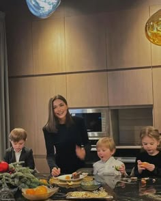 a woman standing in front of a kitchen counter with three children sitting at the table