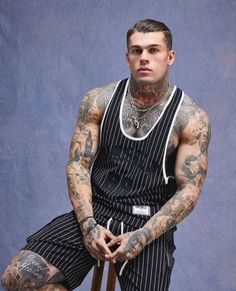 a man with tattoos sitting down wearing a black and white striped dress, holding his hands in his pockets