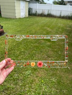 a hand holding up a glass frame with flowers on it in the grass next to a house