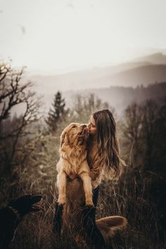 a woman sitting in the grass with her dog and kissing her face while she is surrounded by other dogs