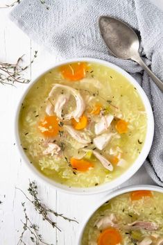 two bowls of chicken and rice soup on a white table with silver spoons next to it