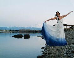 a woman in a white and blue dress standing on rocks near the water with her arms outstretched