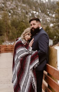 a man and woman standing next to each other on a wooden bridge with snow covered mountains in the background