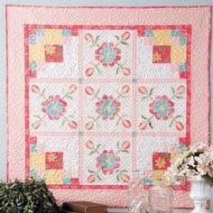 a pink and yellow quilt hanging on the wall next to a vase with flowers in it