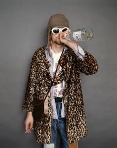 a man wearing sunglasses and a leopard print coat drinking from a water bottle while standing in front of a gray wall