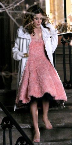 a woman in a pink dress and white coat standing on steps with her hands behind her head