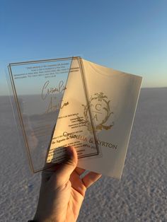 a person holding up two wedding cards in the air with sand and blue sky behind them