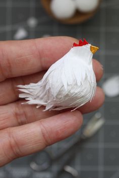 a hand holding a small white chicken in it's left hand and another cupcake on the table behind it