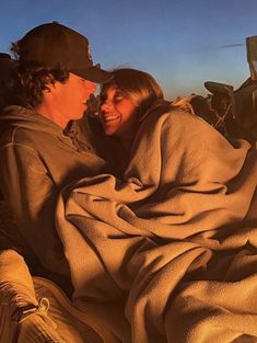 a man and woman wrapped in blankets sitting on the back of a truck together, smiling at each other