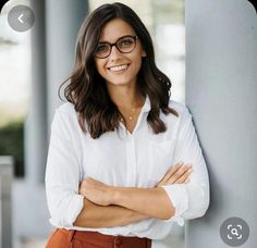 a woman wearing glasses leaning against a pillar with her arms crossed and looking at the camera